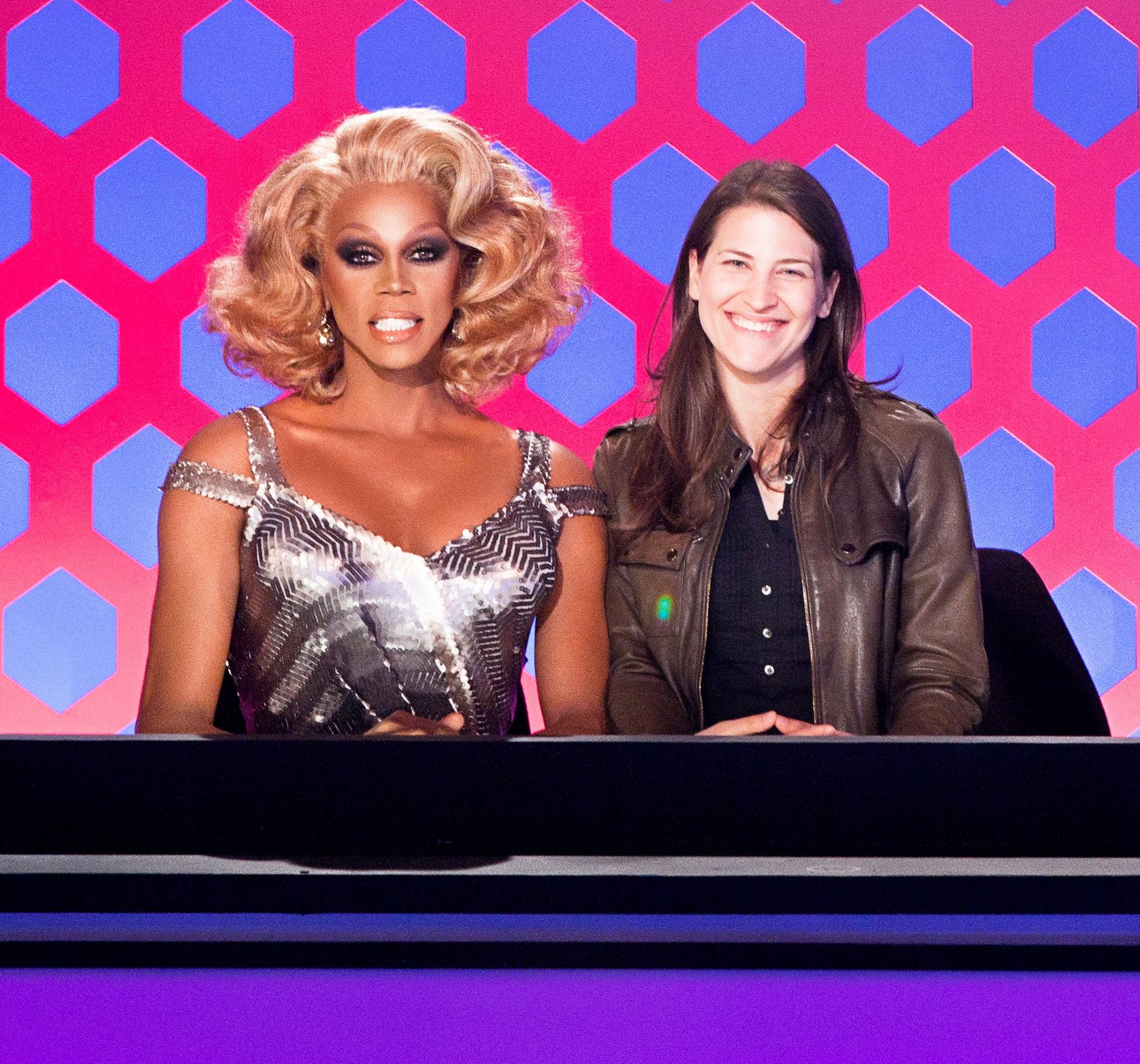 A photograph of drag queen RuPaul, in blond wig and a silver, sparkling dress with two straps on each shoulder, next to the author, who presents as female in long brown hair and a leather jacket over a fitted black shirt with buttons. They are sitting shoulder to shoulder behind a black counter on top of a purple base, the judges’ panel on RuPaul’s Drag Race; behind them is a wall with magenta hexagons on a blue background.