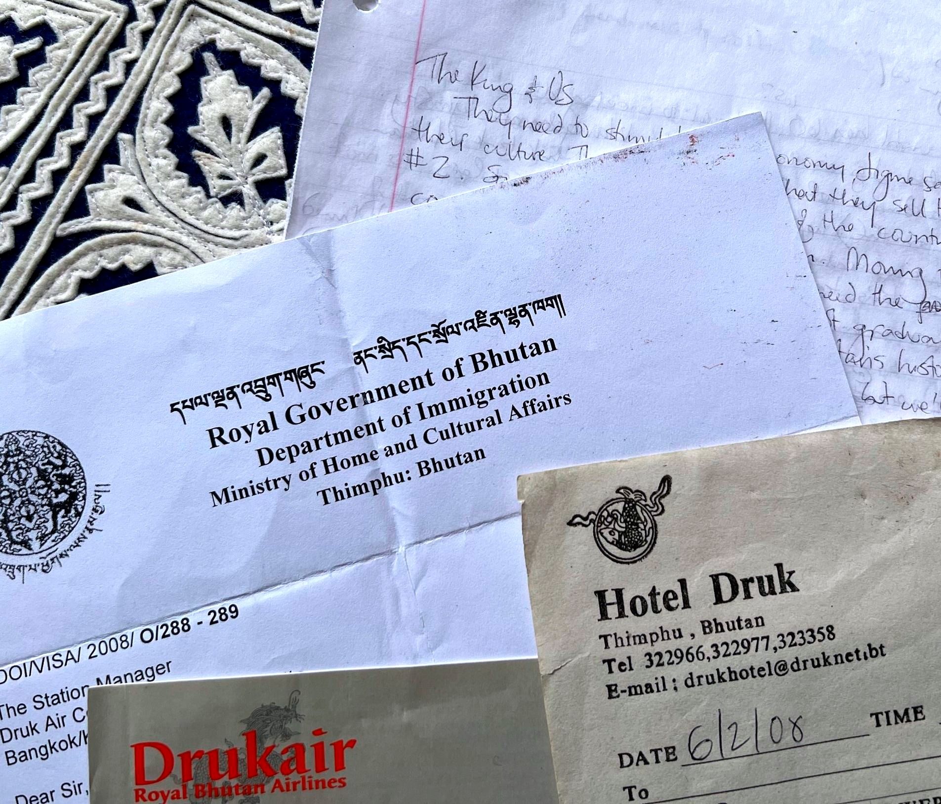 Four documents arrayed against a background of royal-blue-and-white fabric in the lefthand corner. The top right document looks like handwriting on notebook paper, with the heading “The King and Us”; below and to the left of that is a printed document that headed with “Royal Government in Bhutan Department of Immigration” in both Bhutanese and English and appears to be a visa; to the bottom and right of that, a paper headed with the words “Hotel Drunk” and contact information in Thimpu, Bhutan, and the handwritten date 6/02/08; at the bottom of the photo is a darker brown piece of paper that says “Drukair, Royal Bhutan Airlines” in orange text.