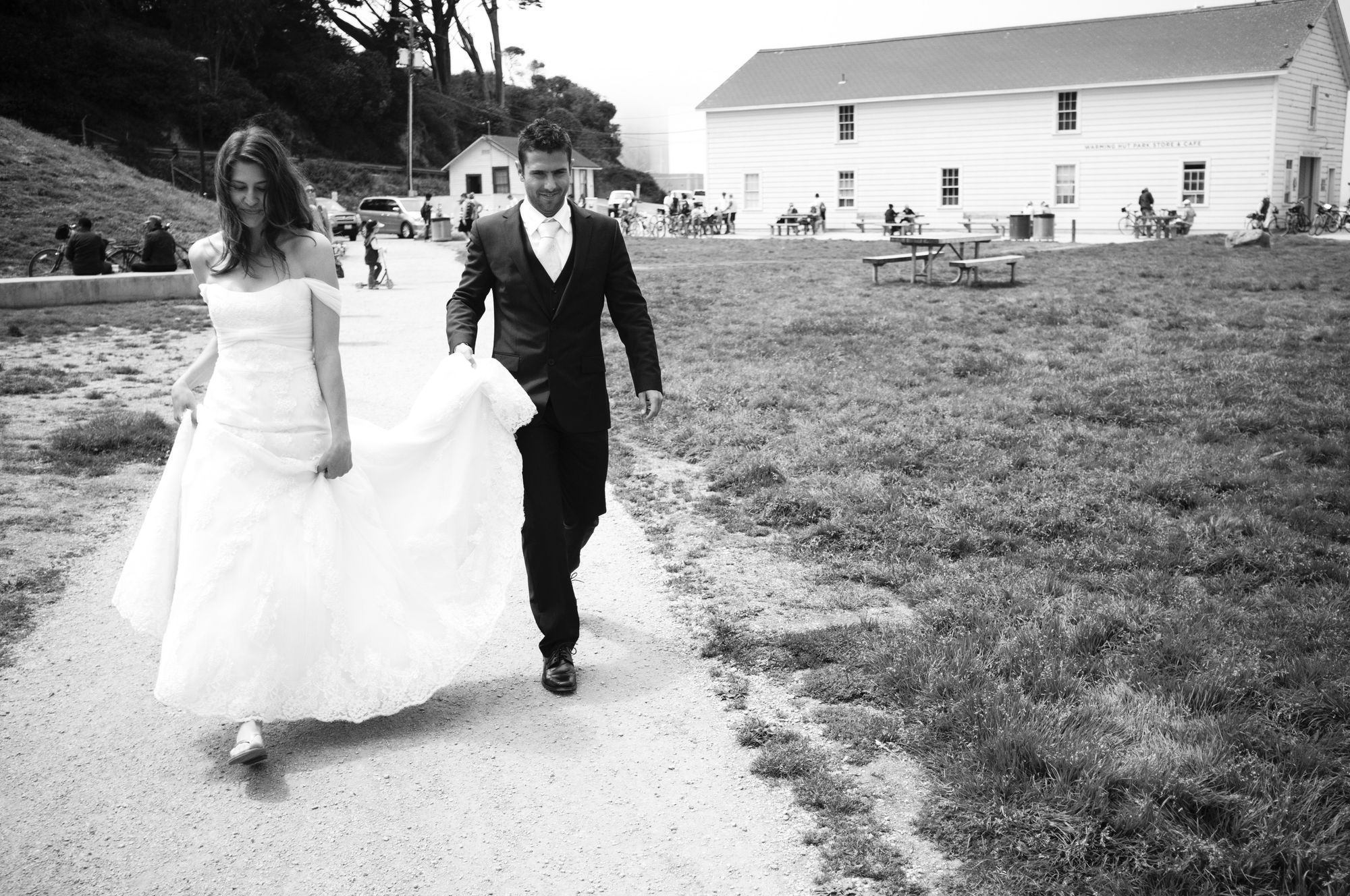 Photo: A couple on their wedding day. The person on the left is the author, female presenting, long brown hair, long white dress with a lace skirt and off-the-shoulder bodice; the man on the right is holding the train as they walk together down a dirt path beset by grass with some picnic tables and plain off-white buildings in the background. The groom on the right is wearing a black suit with a white shirt and cream-colored tie with shiny black shoes. 