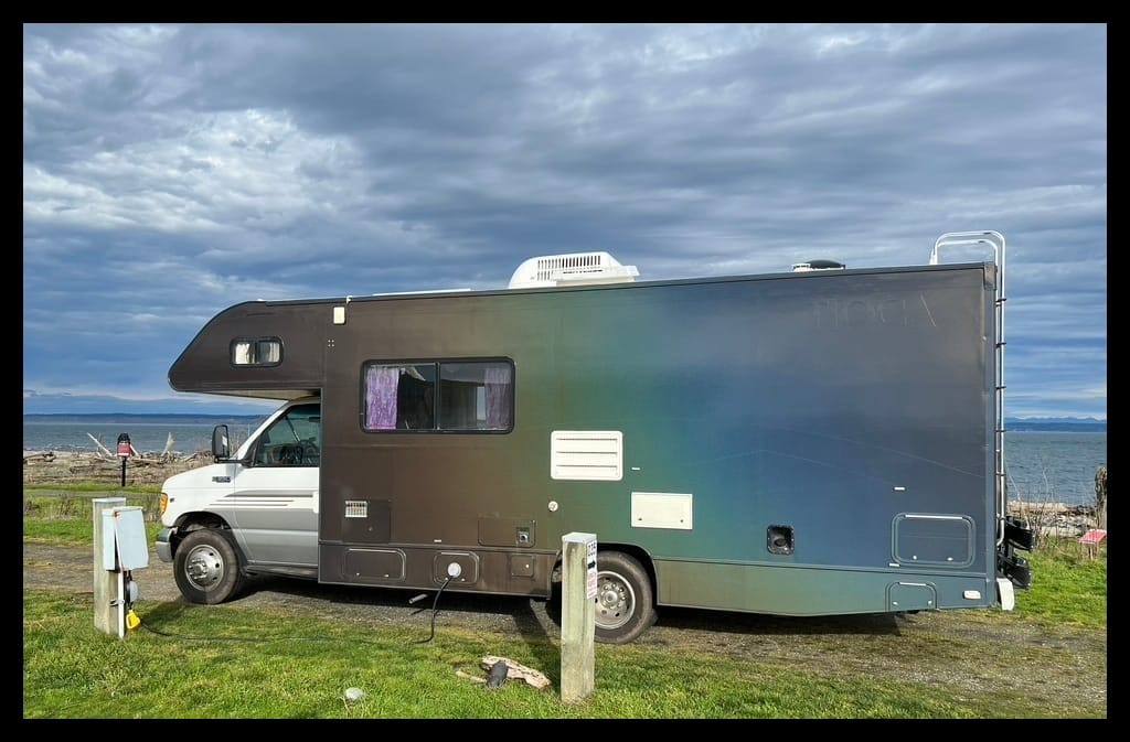 An RV covered in a prismatic material that shines with the colors of a prism along its side, starting with dark rust at the front and fading to orange, to yellow, to green, to blue, to purple at the back of the vehicle. The RV is parked on worn green grass, with flat blue ocean in the background and a cloudy blue sky above. 