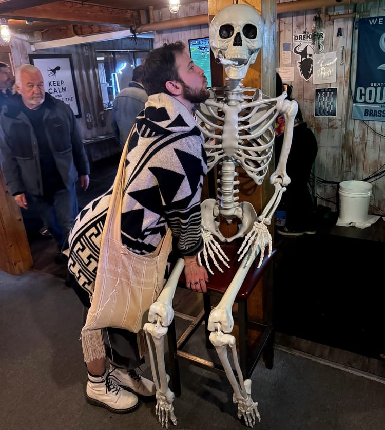The author leans into a life-sized skeleton tacked against a wooden post in a bar, making a kissing face.