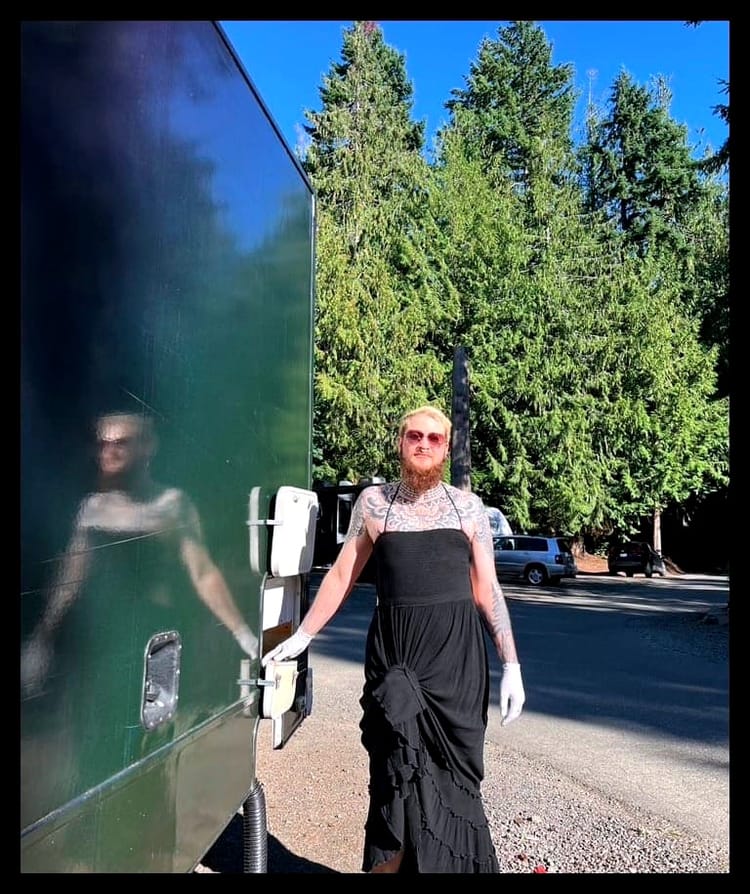 A bearded man in a long black dress stands in front of evergreens next to an RV with a black hose coming out of the bottom.