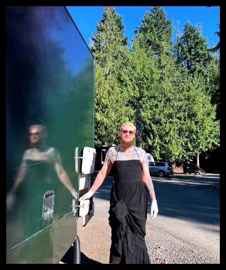 A bearded man in a long black dress stands in front of evergreens next to an RV with a black hose coming out of the bottom.